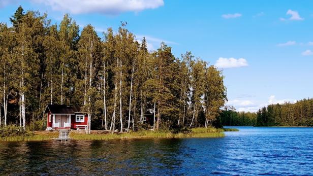 Ein kleines, rotes Haus mit weißen Akzenten steht am Ufer eines Sees, umgeben von Bäumen.