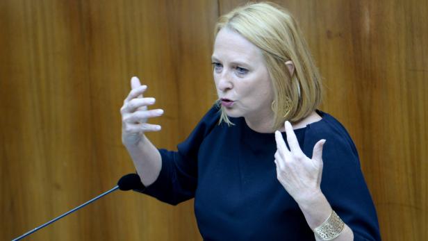 Eine blonde Frau gestikuliert während einer Rede vor einem hölzernen Hintergrund.