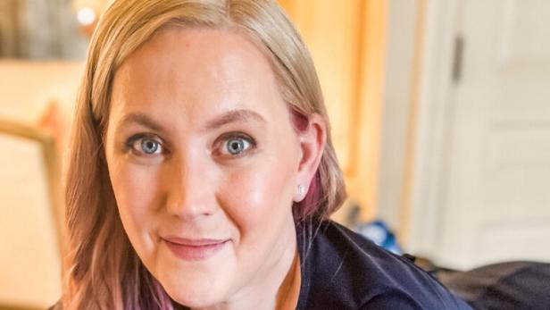 Eine Frau mit blonden und lila Haaren liegt entspannt auf einem Sofa und schaut freundlich in die Kamera.