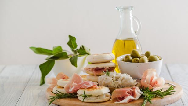Ein Holzteller präsentiert Oliven, Öl, Brötchen mit Schinken und Kräutern.