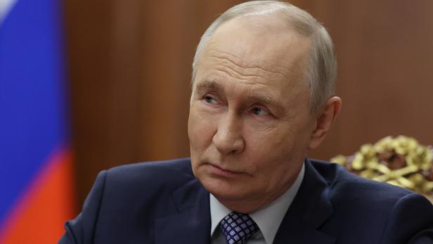 Ein Mann im Anzug mit ernster Miene sitzt vor einer russischen Flagge und schaut zur Seite.