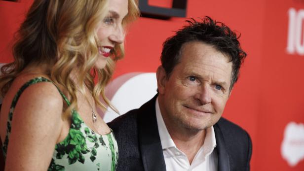 Michael J. Fox im Anzug und seine Frau Tracy Pollan mit blonden Locken lächeln vor rotem Hintergrund.