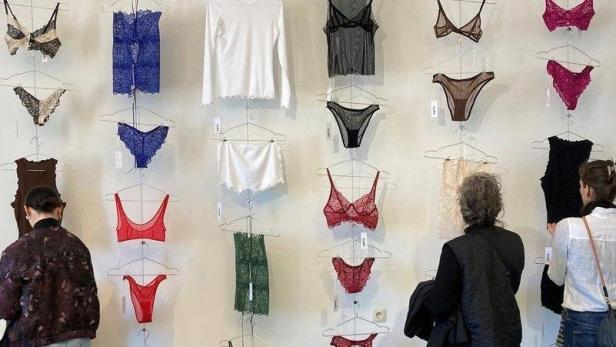 Verschiedene bunte Dessous und Oberteile hängen an einer Wand, während drei Personen davor stehen.
