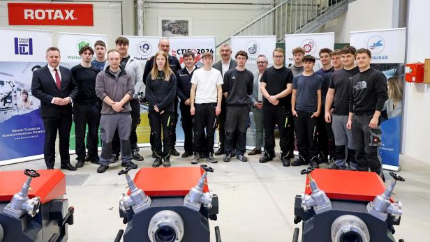 Eine Gruppe von jungen Erwachsenen und mehreren Erwachsenen steht in einer Werkhalle vor technischen Geräten und Informationsplakaten.