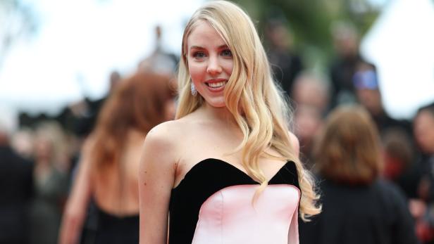 Eine lächelnde Frau mit blonden, gewellten Haaren trägt ein trägerloses Kleid mit schwarzem und rosa Stoff.