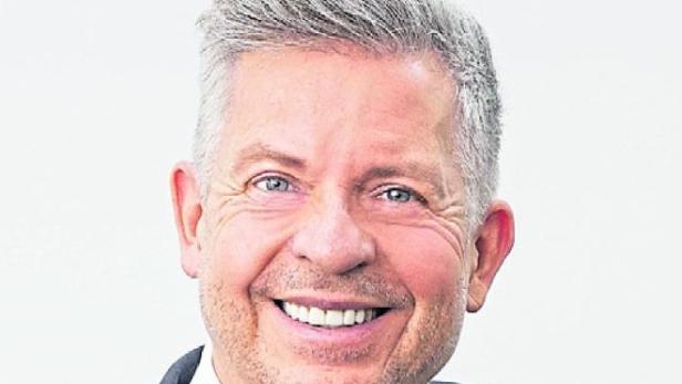 Ein Mann im Anzug mit Krawatte lächelt und steht an einem Treppengeländer vor hellem Hintergrund.