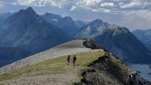 Zwei Wanderer gehen auf einem schmalen Grat mit steilen Abhängen und Bergpanorama.