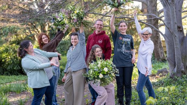 Sieben fröhliche Menschen posieren im Freien, einige halten große Blumensträuße in die Luft.