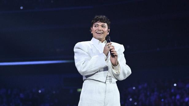 Eine Person in weißem Anzug hält ein Mikrofon und lächelt auf einer Bühne vor großem Publikum.