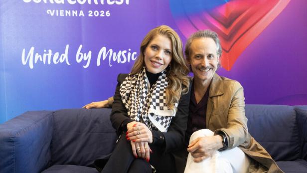 Zwei Personen sitzen vor einem Eurovision Song Contest 2026 Hintergrund auf einem Sofa.