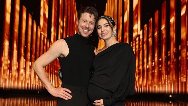 Renata und Valentin Lusin posieren lächelnd zusammen, Renata hält ihre schwangere Bauch, vor einer goldenen Bühnenkulisse.