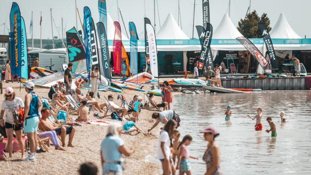 Viele Menschen entspannen am Strand, spielen im Wasser und bunte Fahnen sowie Zelte stehen im Hintergrund.