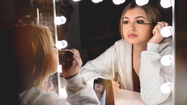 Junge Frau trägt vor einem beleuchteten Spiegel Mascara auf und filmt sich dabei mit ihrem Smartphone.