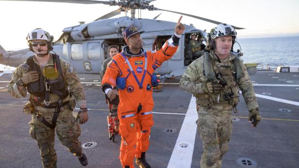 Ein Astronaut im orangefarbenen Raumanzug wird von Soldaten auf dem Deck eines Schiffes mit Hubschrauber begleitet.