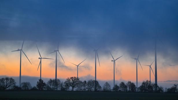 Windräder stehen in einer nebligen Landschaft vor einem Sonnenuntergang, Bäume sind im Vordergrund zu sehen.