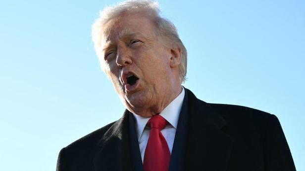 Donald Trump spricht mit offenem Mund vor blauem Himmel, gekleidet in einen dunklen Mantel und roter Krawatte.