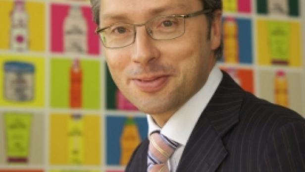 Ein Mann mit Brille und Anzug steht vor einer Wand mit bunten Produktabbildungen.