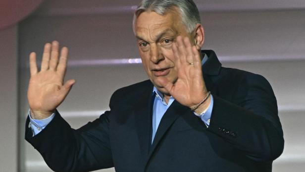 Viktor Orbán winkt mit erhobenen Händen in einem dunklen Anzug.