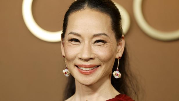 Lucy Liu lächelt mit Ohrringen vor einem braunen Hintergrund.