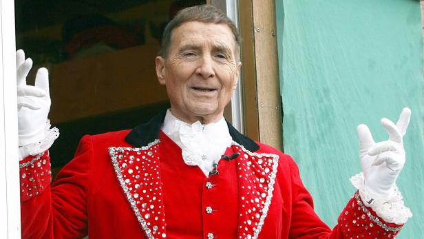 Freddy Quinn trägt einen roten, mit Strass besetzten Frack und weiße Handschuhe und lächelt mit erhobenen Händen.