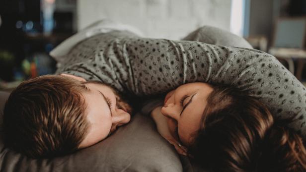 Zwei Menschen liegen eng umarmt auf einem Kissen und schlafen mit geschlossenen Augen nebeneinander.