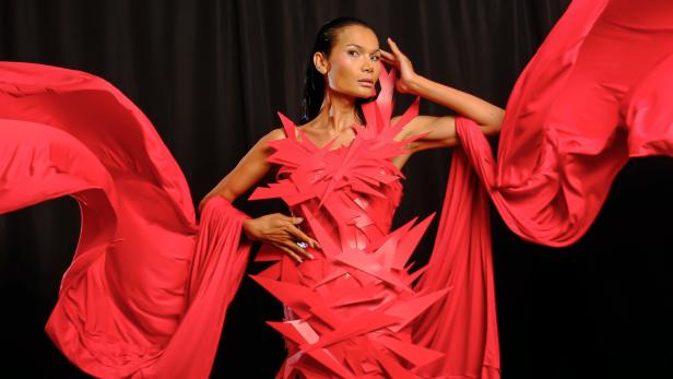 Eine Frau posiert in einem roten Kleid mit zackigen Details und wehenden Stoffbahnen vor schwarzem Hintergrund.