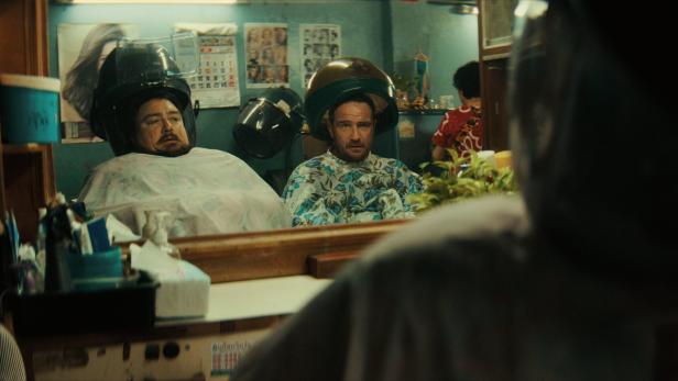 Zwei Männer sitzen unter Haartrocknern in einem Friseursalon und schauen in den Spiegel.