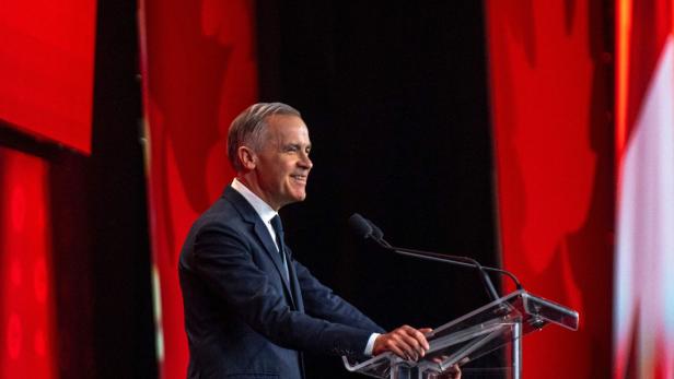 Ein Mann im Anzug spricht lächelnd an einem Rednerpult vor rotem Hintergrund mit Ahornblattmotiv.