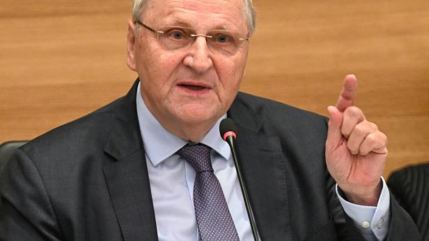 Ein älterer Mann im Anzug spricht am Mikrofon und hebt den rechten Zeigefinger, Holzwand im Hintergrund.