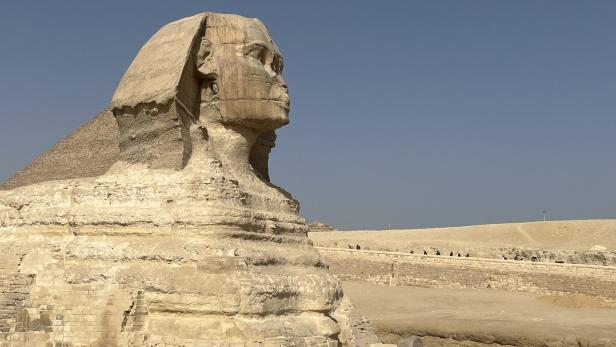Die Große Sphinx von Gizeh mit Löwenkörper und Menschenkopf steht in einer sandigen Wüstenlandschaft.