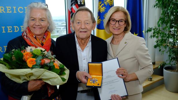 Drei Personen posieren mit einer Auszeichnung und einem Blumenstrauß vor Flaggen.