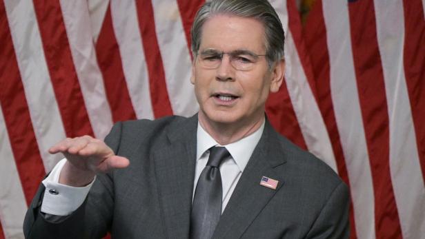 Ein Mann im Anzug mit US-Flaggen-Anstecker spricht gestikulierend vor mehreren amerikanischen Flaggen.