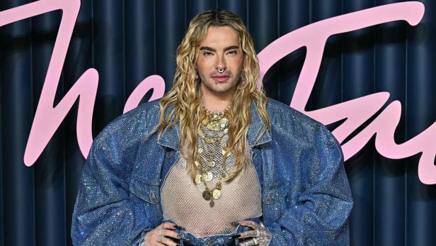 Bill Kaulitz mit blonden, gewellten Haaren, Nasenring und vielen Halsketten trägt eine glitzernde Jeansjacke.