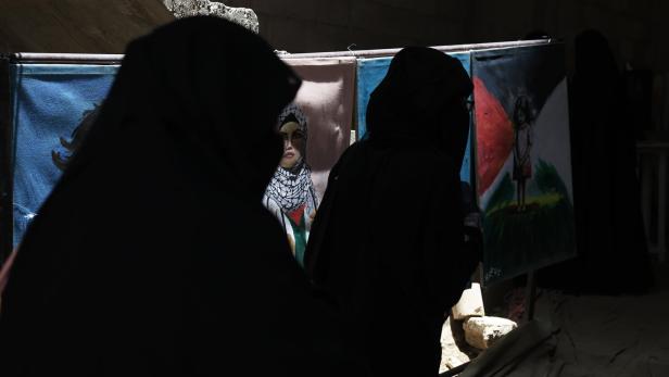 Zwei Frauen in dunkler Kleidung stehen vor Gemälden, darunter Porträts und ein Kind vor einer Flagge.