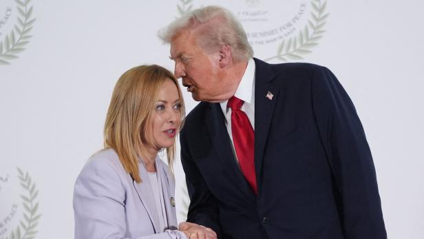 Donald Trump schüttelt Giorgia Meloni die Hand und beugt sich ihr zu, während sie sich vor einem hellen Hintergrund mit Lorbeerkranz-Symbolen ansehen.