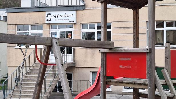 Ein Spielplatz mit Klettergerüst und Rutsche steht vor einem Gebäude mit dem Schild „JA Don Bosco Jugendzentrum Amstetten“.