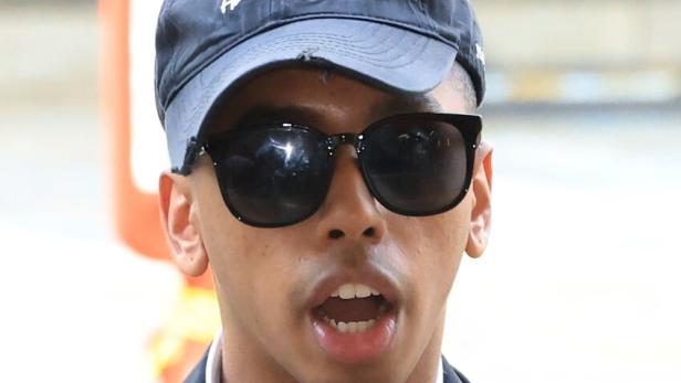 Ein Mann mit Sonnenbrille und schwarzer Kappe trägt Anzug, Rucksack und mehrere Mikrofone am Revers.