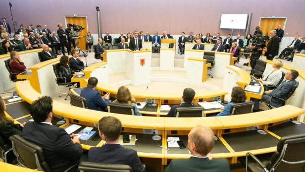 Mehrere Personen sitzen in einem modernen Plenarsaal, während eine Person am Rednerpult spricht.