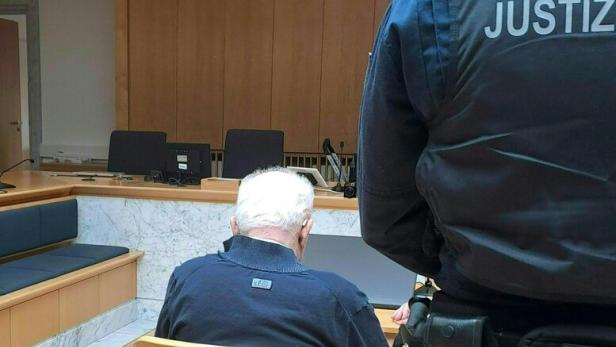 Ein Mann mit weißem Haar sitzt in einem Gerichtssaal, bewacht von einem Justizbeamten.