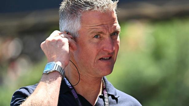 Ralf Schumacher mit grauem Haar und blauem Hemd hält einen Ohrhörer ins Ohr.