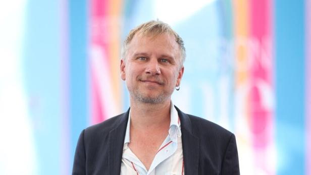 Robert Stadlober mit Sakko und gestreiftem Hemd steht vor einem farbenfrohen Hintergrund.