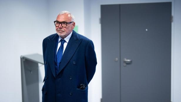 Älterer Mann mit Brille, dunklem Mantel und Krawatte vor dunkler Tür.