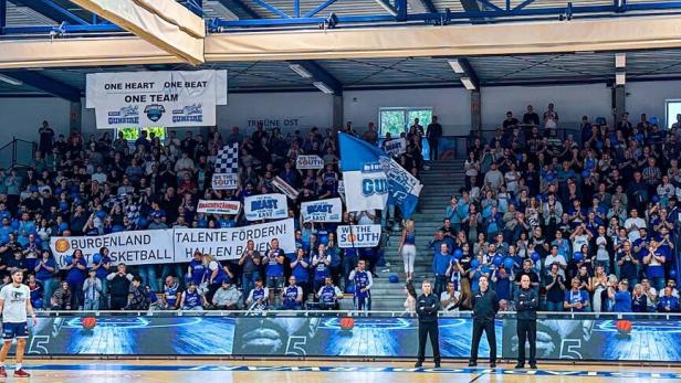 Viele Fans in blauer Kleidung und mit Bannern feuern ein Basketballteam in einer vollen Sporthalle an.
