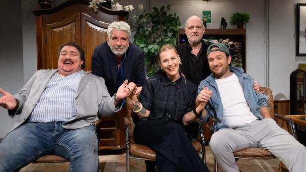 Fünf lachende Personen sitzen und posieren gemeinsam in einem gemütlichen Fernsehstudio.