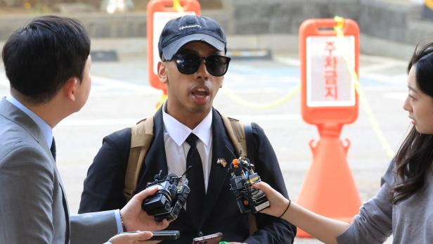 Ein Mann mit Sonnenbrille, Anzug und Basecap wird draußen von zwei Reportern mit Mikrofonen interviewt.