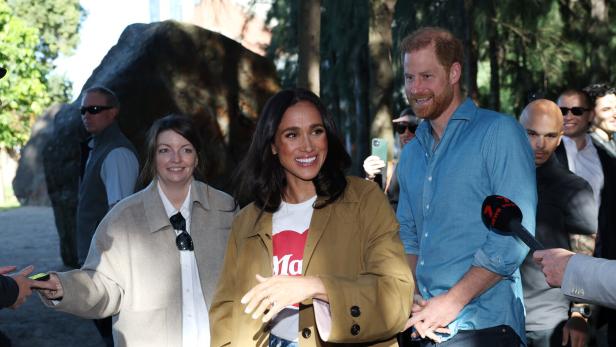 Meghan und Harry lächeln umgeben von Menschen und Mikrofonen im Freien.