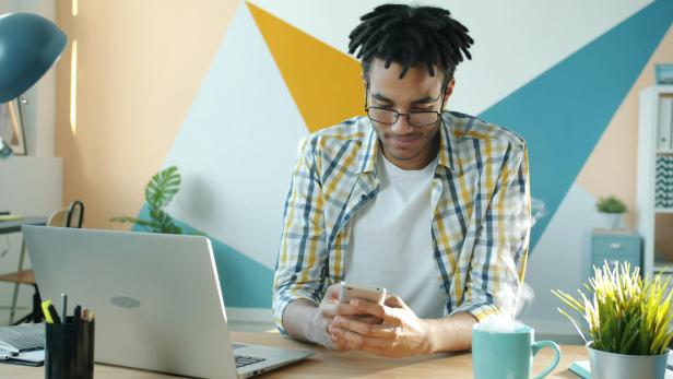 Junger Mann arbeitet am Schreibtisch mit Laptop und Smartphone, daneben eine Tasse Kaffee.