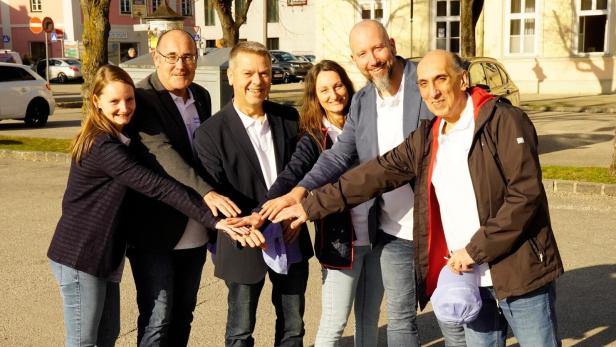 Sechs Erwachsene stehen im Kreis und legen ihre Hände übereinander, alle lächeln und wirken freundlich.