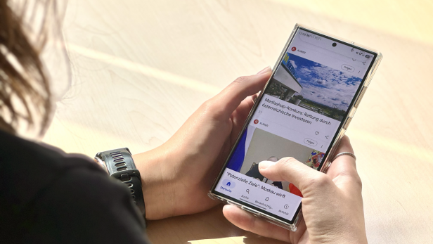 Person hält ein Smartphone und scrollt durch den Google-Discover-Feed mit KURIER-Artikeln.