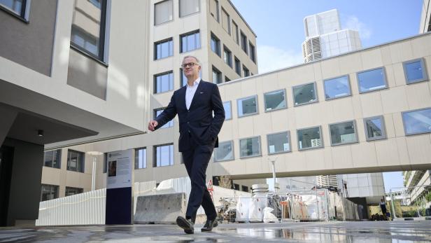 Ein Mann im Anzug geht selbstbewusst durch einen modernen Gebäudekomplex mit Baustellenbereich.